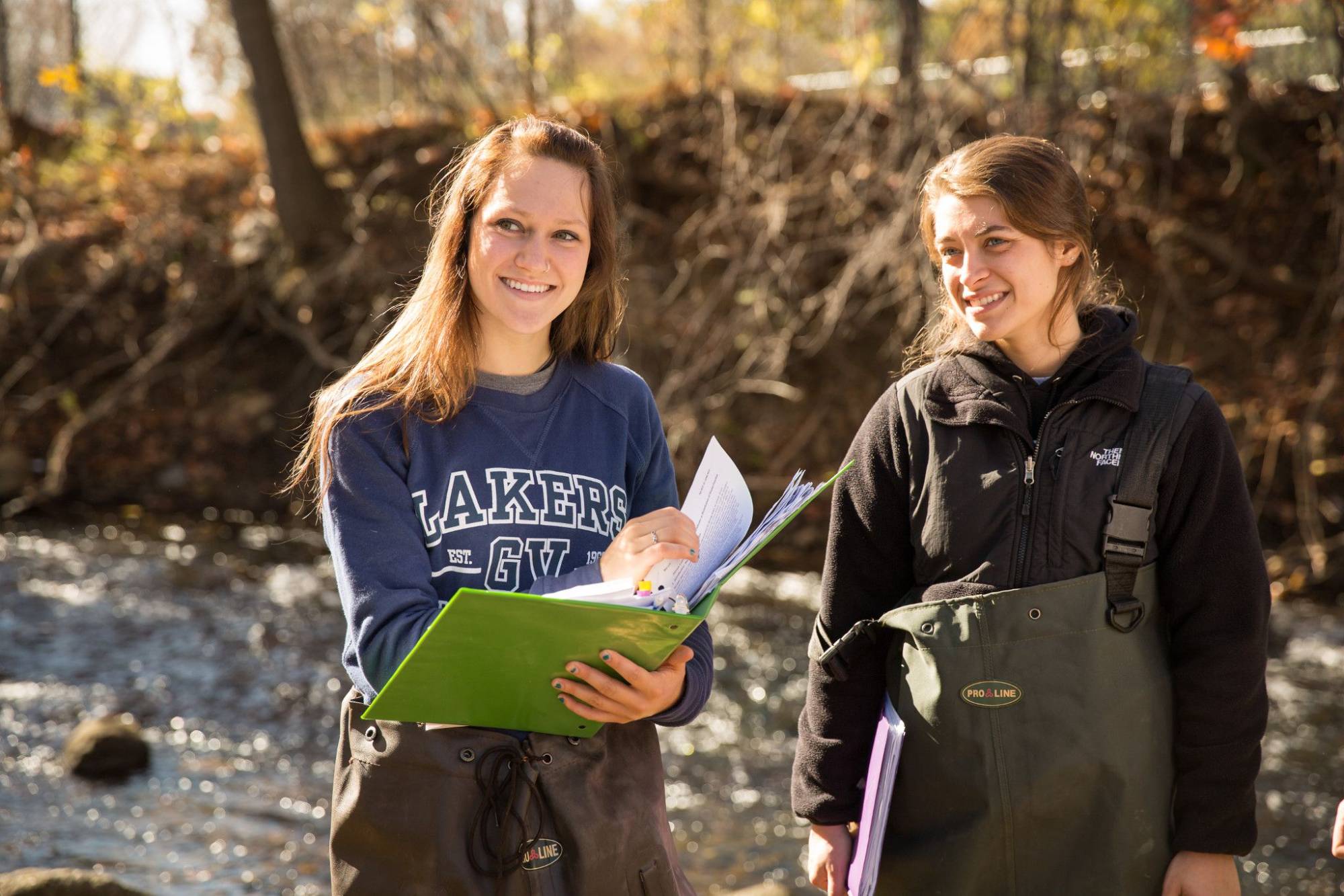 Two young white women on a field experience for a Biology course; they are standing by a river, both are wearing wading gear, and one is holding a large binder.